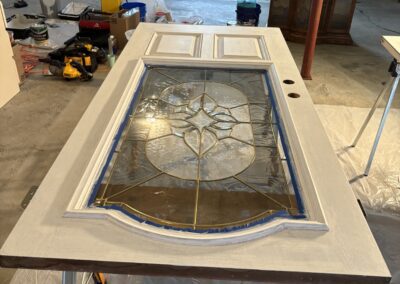 A wooden door with a decorative glass window is laid flat on sawhorses for painting by an Upstate New York painting contractor in a room filled with tools, supplies, and a large window. The door is partially painted white.