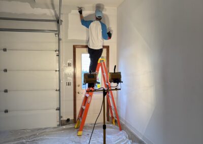 A painter Upstate New York stands on a ladder, painting the upper wall of a garage. Plastic sheeting covers the floor, and a work light brightens the workspace—showcasing professional interior painting Upstate New York.