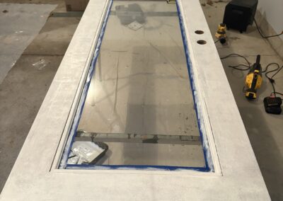 A glass-paneled door lies flat on a table in a garage, being painted white by a residential painting Upstate New York expert. Painting tools, a drill, bucket with rags, and sink are visible; the window is taped off for painting.